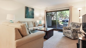 A living room with a sofa, armchair, and coffee table. There is a television and a lamp on a stand. A large window with curtains is visible. - Ocean One 120 - Beachside Condo (Hilton Head Island)