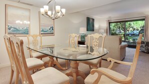 A dining area with a glass table surrounded by wooden chairs with cushioned seats. There is a chandelier above the table and artwork on the walls. - Ocean One 120 - Beachside Condo (Hilton Head Island)