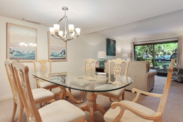 A dining area with a glass table surrounded by wooden chairs with cushioned seats. There is a chandelier above the table and artwork on the walls.