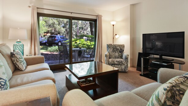A living room with a sofa, armchair, and a TV. There is a glass table and a lamp. The room has a large window with curtains. - Ocean One 120 - Beachside Condo (Hilton Head Island)