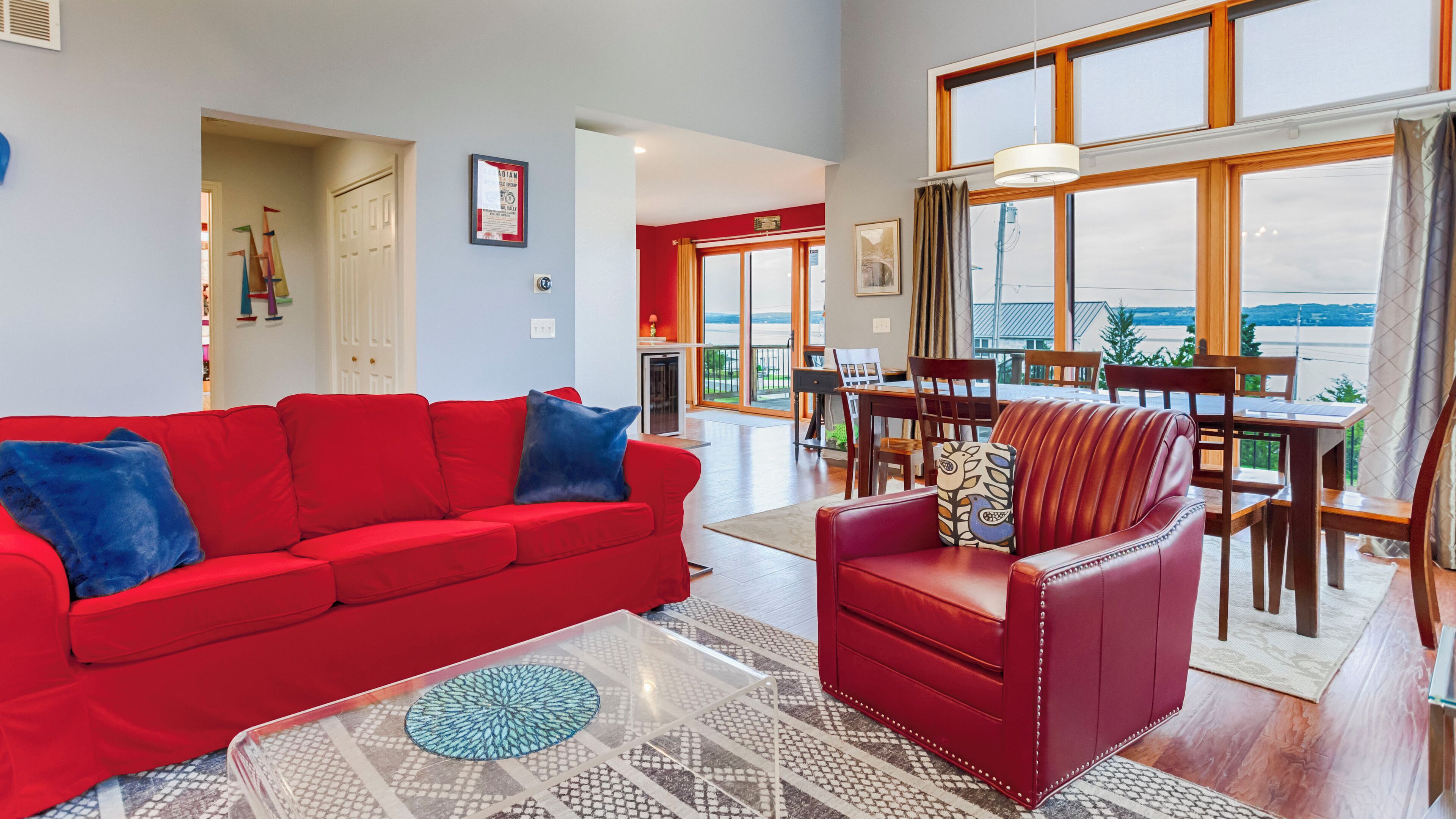 Un salon spacieux avec un canapé rouge et un fauteuil en cuir rouge, un coin repas avec des meubles en bois, et de grandes fenêtres offrant une vue sur l’extérieur.