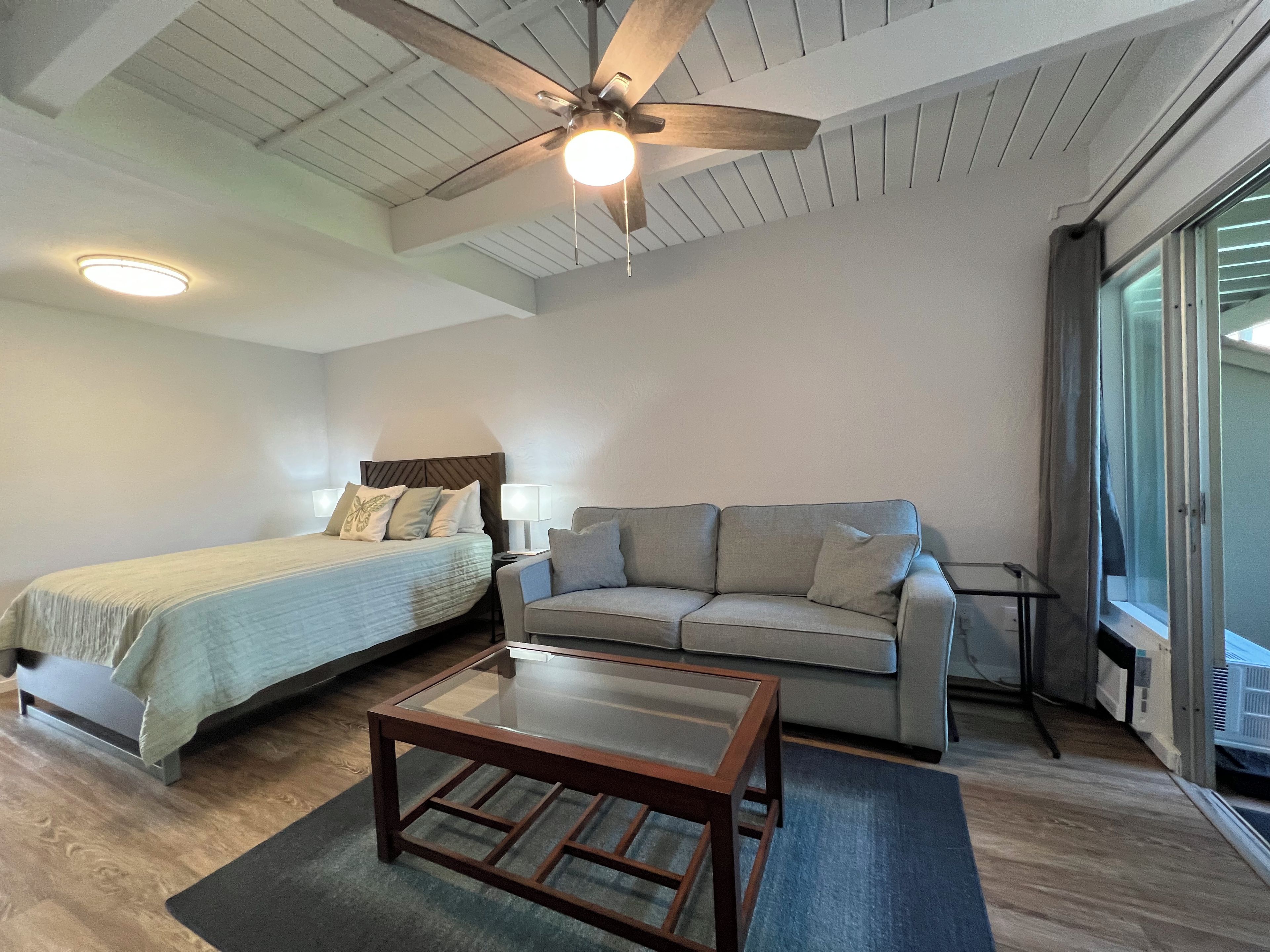 A hotel room with a bed, a sofa, a coffee table, a ceiling fan, and a window.