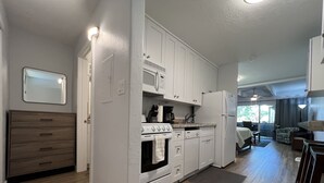 A modern kitchen with white cabinets, a stove, and a refrigerator. There is a wooden cabinet and a dining area with a table and chairs. - Kihei Bay Surf #123 A/C, Wifi, Near Beach, Great Rates, Shared Hot Tub (Kihei)