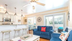 A bright living room with a blue sofa, white coffee table, and a ceiling fan. There is a kitchen area with white cabinets and a microwave. - Captains Watch 11 | Rooftop Pool, 1 Block to Beach (Tybee Island)
