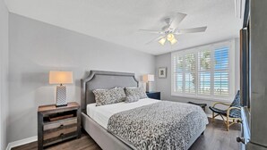 Ein Schlafzimmer mit einem Bett, Nachttischen, einem Deckenventilator, einem Fenster mit Jalousien und einem Sessel.