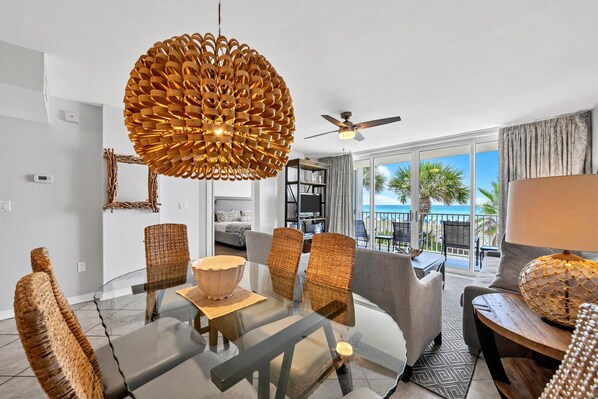 A modern living room with a glass dining table, wicker chairs, a unique spherical chandelier, and a view of the beach through large windows.