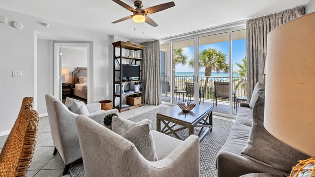 Ein geräumiges Wohnzimmer mit Deckenventilator, Fernseher und Bücherregal. Ein Balkon bietet Blick auf den Strand und Palmen.