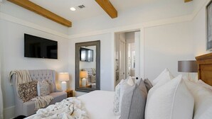 A bedroom with a large bed, a TV mounted on the wall, a mirror, and a wooden ceiling beam. - Driftwood - Vacation Home Near the Beach with a Large Outdoor Area (Santa Rosa Beach)