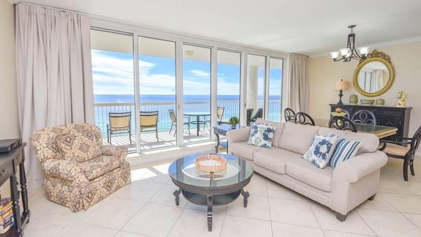 A living room with a sofa, armchair, and coffee table, a dining area with chairs and a table, and a large sliding glass door leading to a balcony with a view of the ocean.