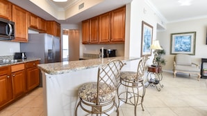 A kitchen with wooden cabinets, a stainless steel refrigerator, granite countertops, and a bar area with two wicker chairs.