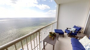 Een balkon met uitzicht op zee, twee blauwe loungestoelen, een tafeltje met een vaas en een kopje, en een hekwerk.