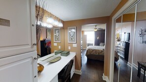 Une chambre d’hôtel avec un lit, un bureau avec un ordinateur, une commode et un placard. Des tableaux encadrés ornent le mur et un ventilateur de plafond est installé dans un coin.