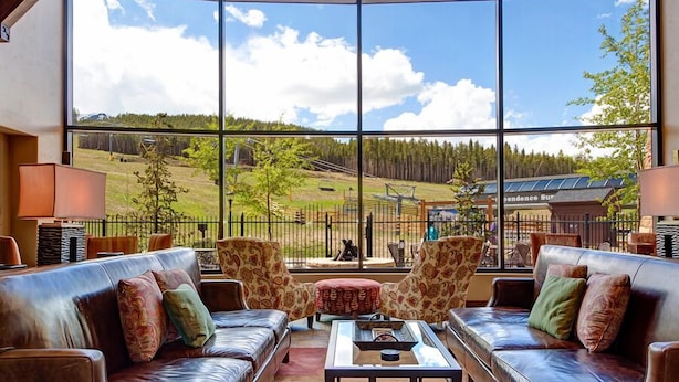 A spacious living area with leather sofas and patterned armchairs, large windows offering a view of a green landscape and a building with solar panels. - Crystal Peak Explorer Condo: Ski-In, Ski-Out Luxury (Breckenridge)