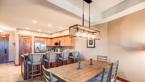A modern kitchen with wooden cabinets, a stainless steel refrigerator, and a dining area with a wooden table and chairs. - Crystal Peak Explorer Condo: Ski-In, Ski-Out Luxury (Breckenridge)