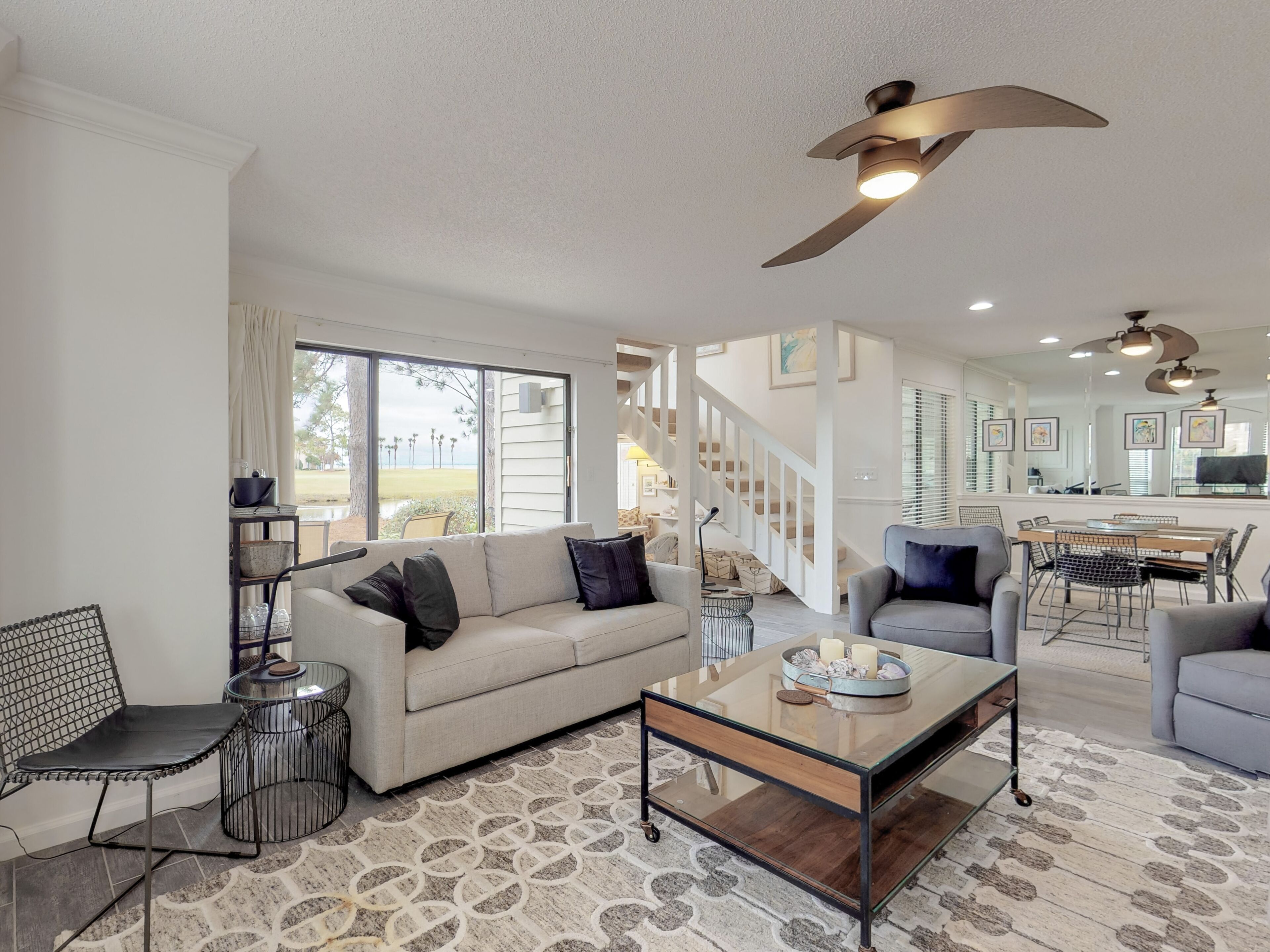 A spacious living room with a sofa, armchairs, a coffee table, and a dining area with a table and chairs. There are ceiling fans and a staircase leading to an upper level.