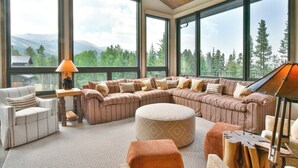 A spacious living room with large windows, a sectional sofa, a round ottoman, and a piano. - Lonestar Lodge: Sweeping Mountain Views, Hot Tub & Patio (Breckenridge)