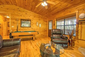 A wooden cabin interior with a pool table, leather chairs, a glass coffee table, and a ceiling fan.