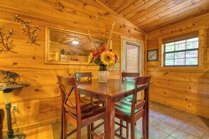 A wooden interior with a dining table, chairs, a vase with flowers, a mirror, and a window.