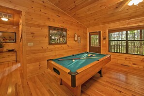 Una habitación de madera con una mesa de billar, paredes de madera y ventanas que dan a un paisaje de árboles.