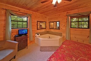 Habitación de cabaña de madera con cama, un pequeño sofá, televisor, bañera y dos ventanas con cortinas.