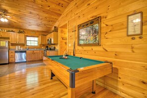 A wooden cabin interior with a pool table, kitchen area, and wooden walls.
