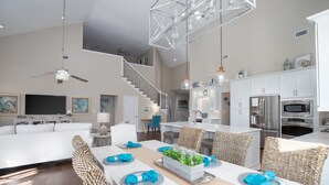 A spacious living area with a dining table set for six, a kitchen with modern appliances, and a staircase leading to an upper level. - 4BR Beach Villa w/ 6-Seater GOLF CART! Near Beach, Pool, Tram, Screened Porch (Miramar Beach)