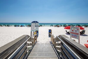 Une promenade en bois menant à une plage où se trouvent des parasols et un panneau indiquant la présence d’un sauveteur.