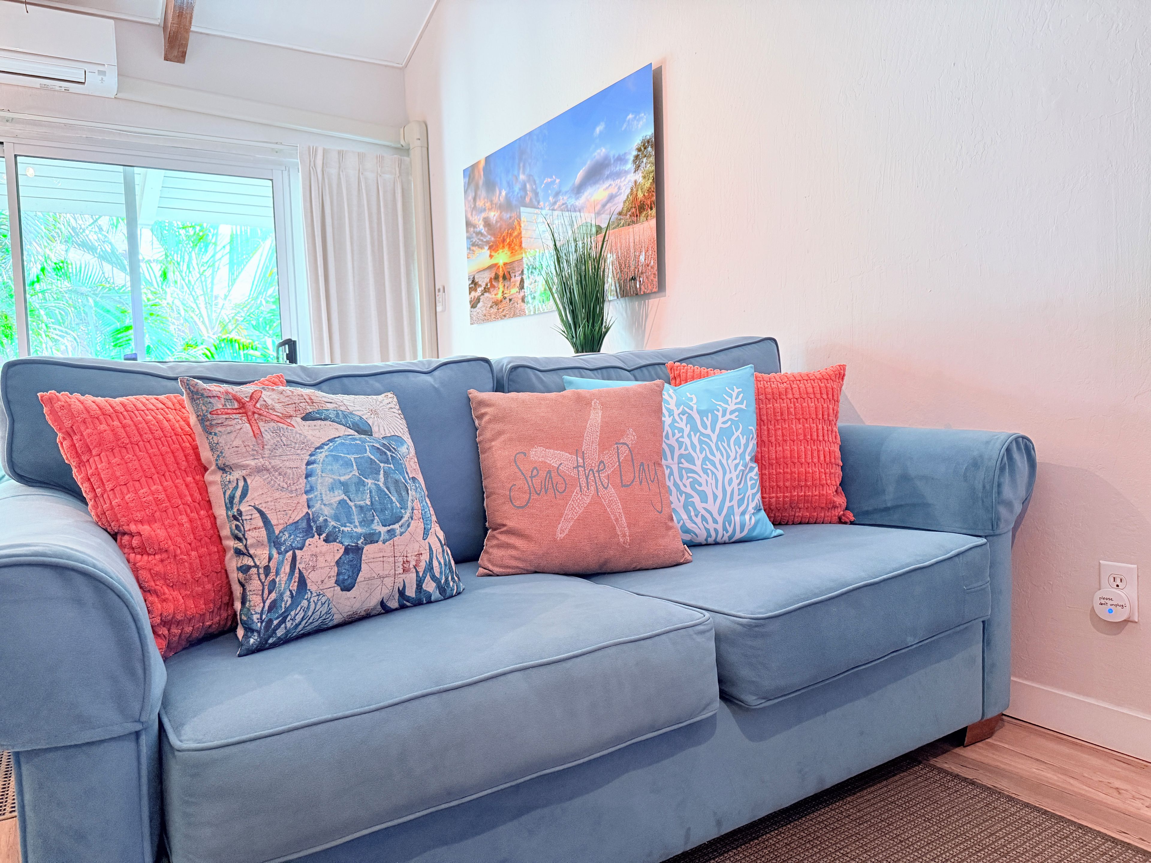 A light blue sofa with four decorative pillows, a window with curtains, and a wall-mounted painting.