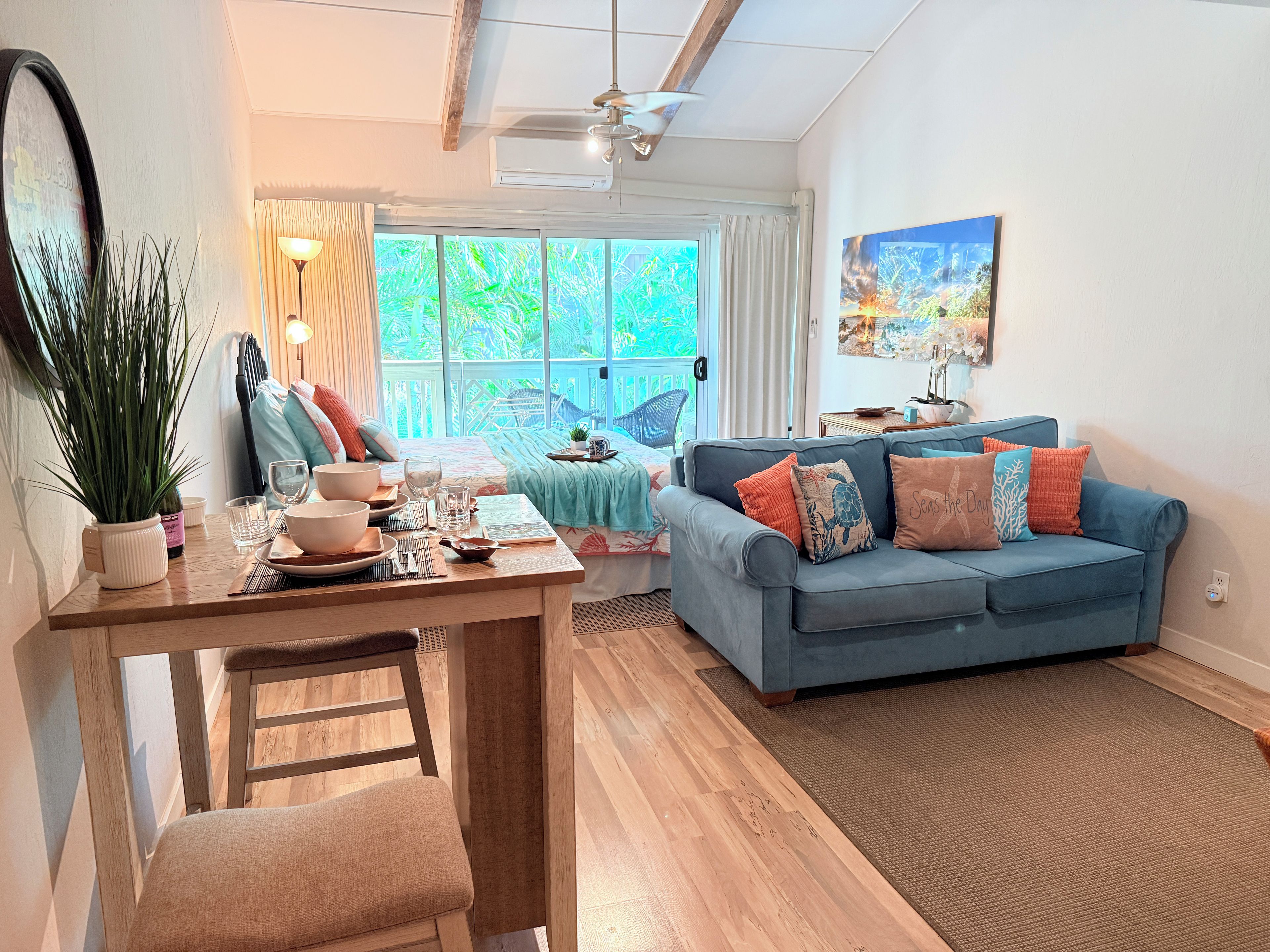 A well-lit living room with a dining area, a blue sofa with colorful cushions, and a large window offering a view of greenery.