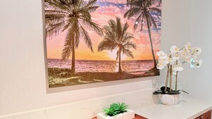 Una pared con una pintura de playa tropical, una orquídea en maceta y una planta pequeña sobre una bandeja de madera.