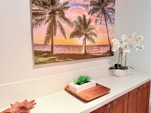 Un muro decorato con un dipinto di una spiaggia tropicale, un'orchidea in vaso e una piccola pianta su un vassoio di legno.