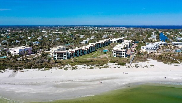 Vista aérea de un hotel frente al mar, con varias construcciones, una playa y un puerto deportivo.