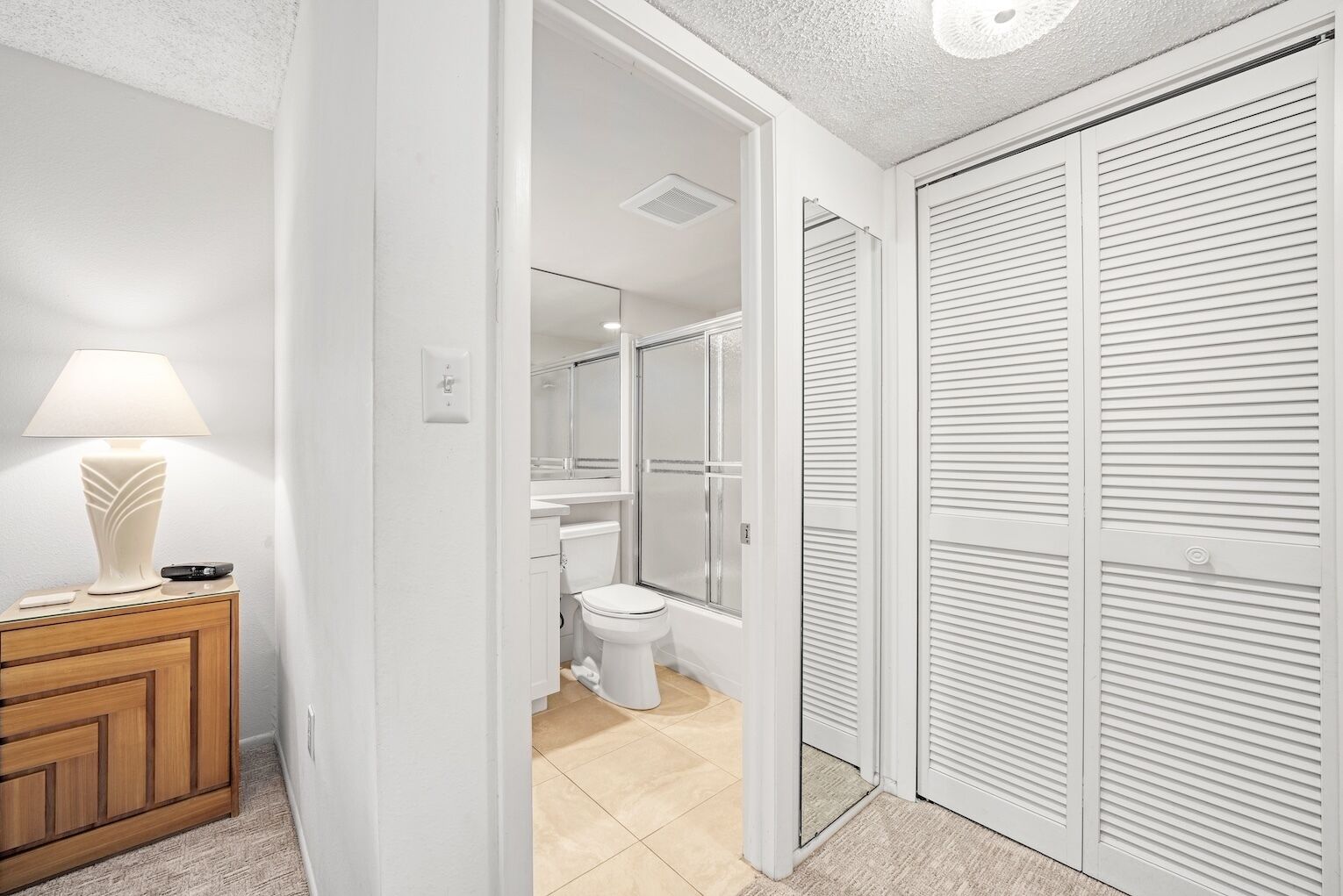 A hallway leading to a bathroom with a toilet, a shower, and a mirror. There is a wooden cabinet with a lamp on it.