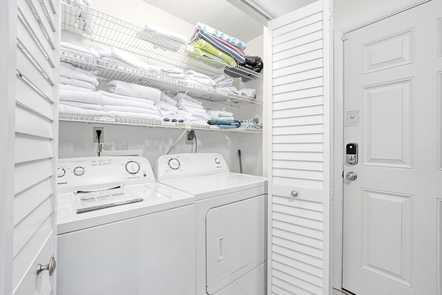 A white laundry room with two washing machines, a dryer, and shelves for folded towels and laundry.