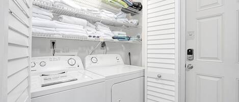 A white laundry room with two washing machines, a dryer, and shelves for folded towels and laundry.