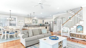 A modern kitchen with white cabinets, a dining area with a white table and chairs, a gray sofa, and a staircase with wooden railings. - Three Sisters | Home with Pool, Patio, Two Porches (Tybee Island)