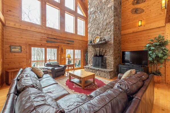 A log cabin interior with a stone fireplace, leather sofas, a television, and large windows. - Luxury Log Chalet w/ Pool & Fireplace (Oakland)