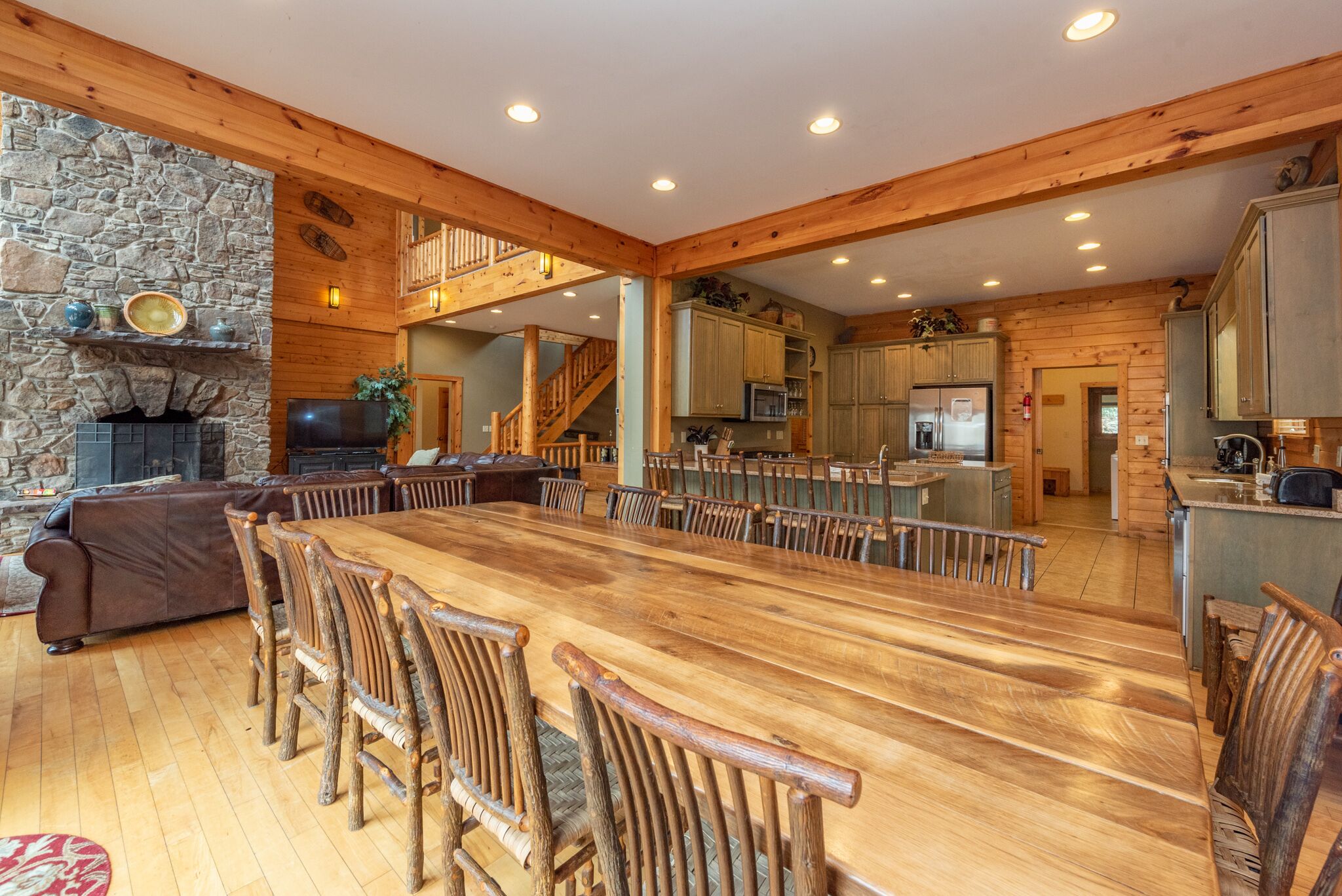 Un vaste espace dédié à la salle à manger, avec une longue table en bois et des chaises en bois au style rustique, une cheminée en pierre et un coin salon confortable avec un canapé en cuir marron.