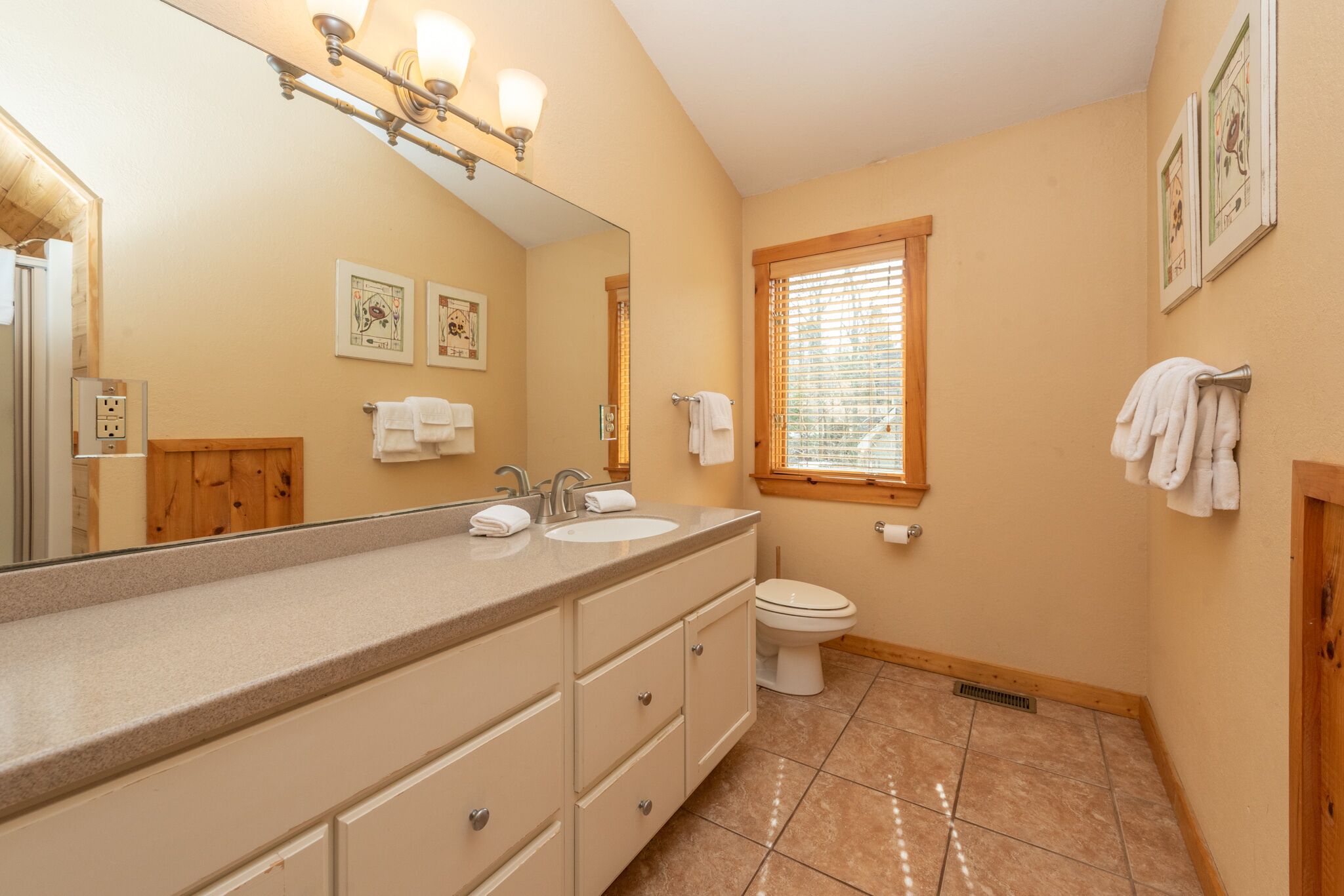 A bathroom with a large mirror, two sinks, a toilet, and a window with blinds.