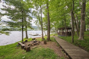 A wooden deck leading to a gazebo by a lake with trees and a fire pit.