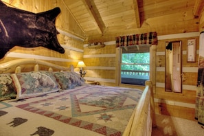 A log cabin bedroom with a large bear head decoration, a quilt with animal prints, and a window with a view of trees.