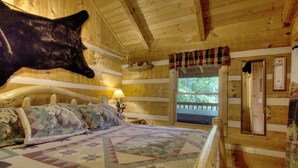 A log cabin bedroom with a large bear head decoration, a quilt with animal prints, and a window with a view of trees.