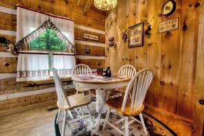 Interior de una cabaña de madera con una mesa redonda para comer, sillas blancas, una ventana con cortinas y un candelabro.
