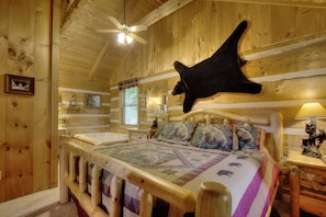 A wooden cabin bedroom with a bed, a mounted bear head, a ceiling fan, and a window with a view of trees.