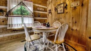 A wooden cabin interior with a round dining table, white chairs, a window with curtains, and a chandelier.