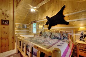 Un dormitorio de cabaña de madera con una cama, una cabeza de oso montada, un ventilador de techo y una ventana con vista a árboles.