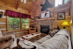 A cozy log cabin interior with a stone fireplace, wooden furniture, and framed pictures on the walls.
