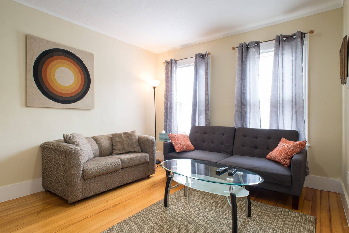 A living room with a brown sofa, a glass coffee table, and a grey sofa with patterned cushions.