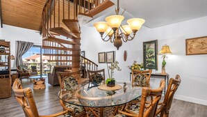 A spacious living area with a glass-top dining table surrounded by wicker chairs, a wooden spiral staircase, and a chandelier.