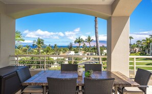 A covered outdoor seating area with a view of the ocean and palm trees.
